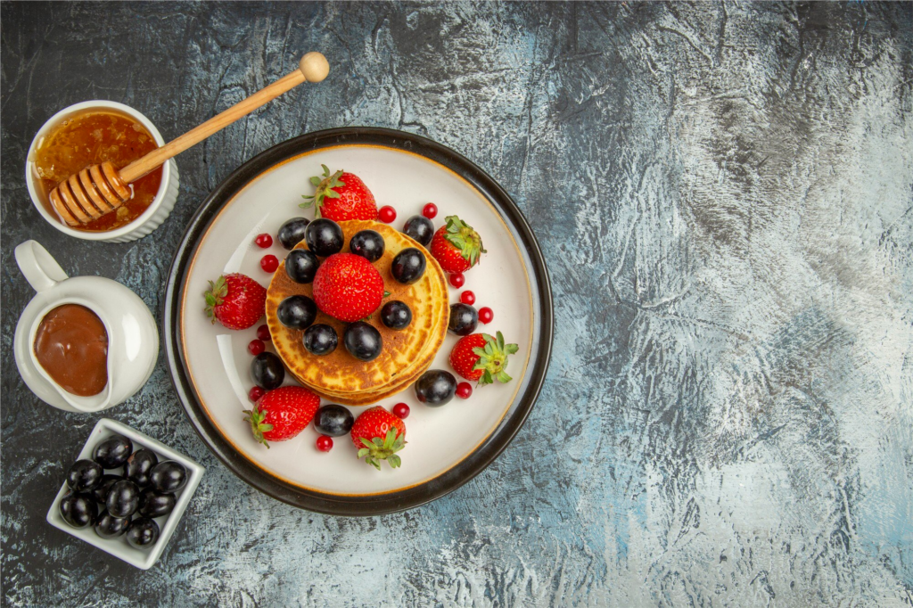 "An appetizing breakfast plate featuring pancakes topped with fresh strawberries, blueberries, and red currants, accompanied by bowls of honey, chocolate sauce, and extra blueberries, arranged on a textured gray-blue background."